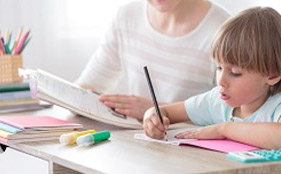 Child writes on paper while unrecognizable adult sits beside them