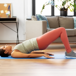 A woman wearing yoga clothing is in her living room, which has a hardwood floor. She is on her back on a yoga mat, doing a bridge exercise — raising her hips into the air while keeping her shoulders and feet flat on the mat. 