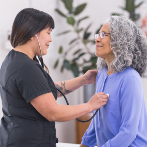 A healthcare provider holds a stethoscope to a patient’s chest as part of a preventative healthcare visit.